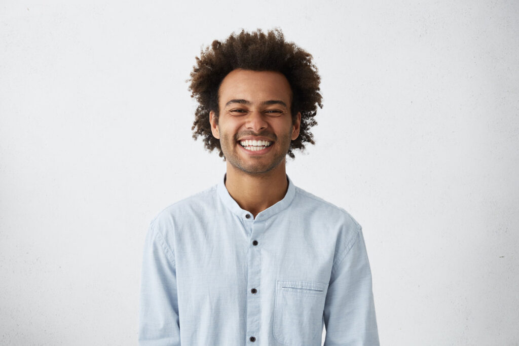 Handsome unshaven young dark-skinned male laughing out loud at funny meme he found on internet, smiling broadly, showing his white straight teeth. Positive human facial expressions and emotions