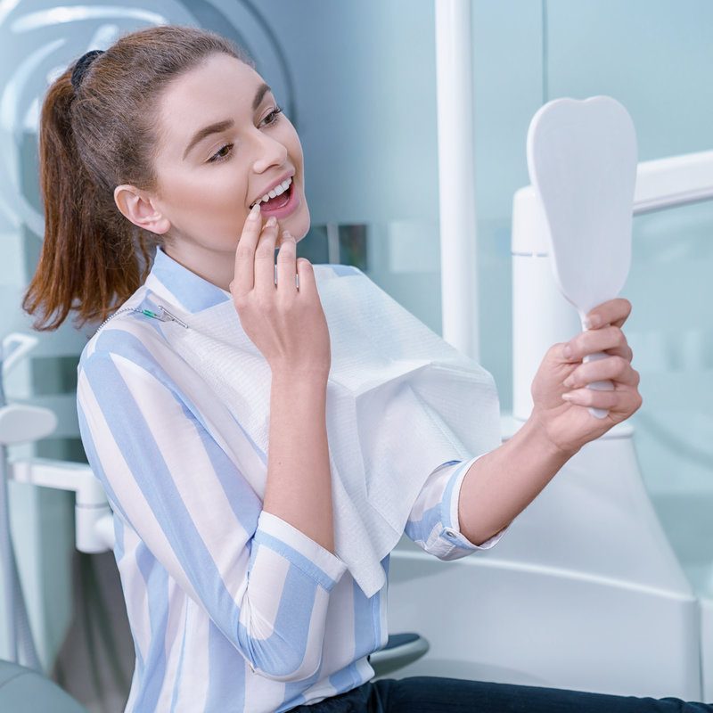 Happy woman satisfied with professional private clinic of dentistry. Charming and adorable woman looking at mirror with opened mouth, smiling. Client holding tooth shaped mirror.